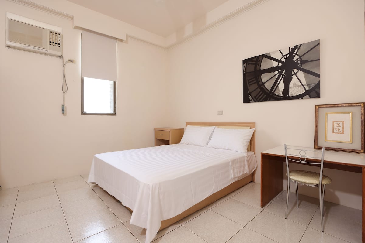 A bedroom with a bed, a desk, a chair, a window, and a wall-mounted air conditioner.