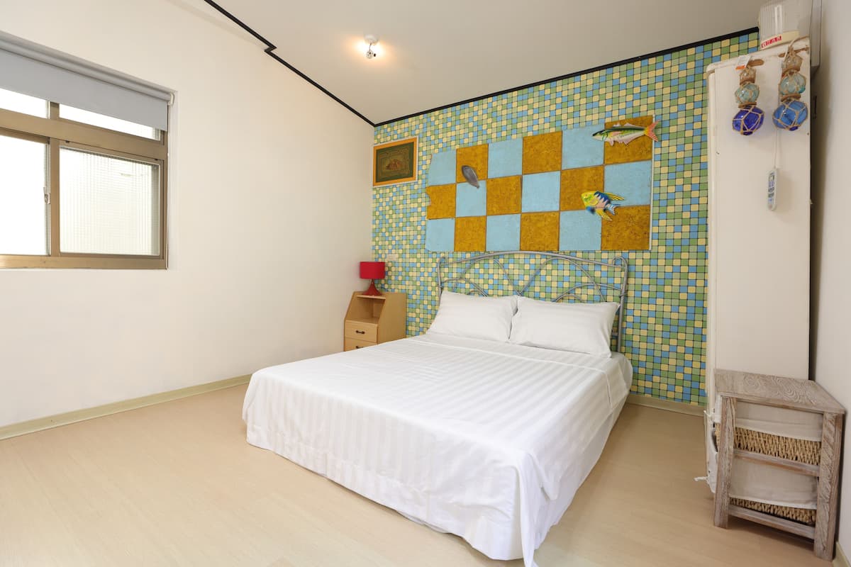 A bedroom with a bed, a window, a wall with a mosaic pattern, and a small wooden nightstand.
