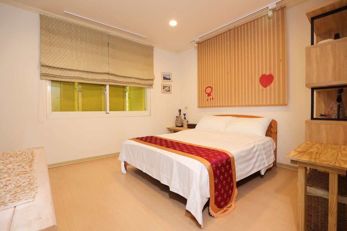 A bedroom with a bed, a nightstand, a wooden shelf, and a window with blinds.