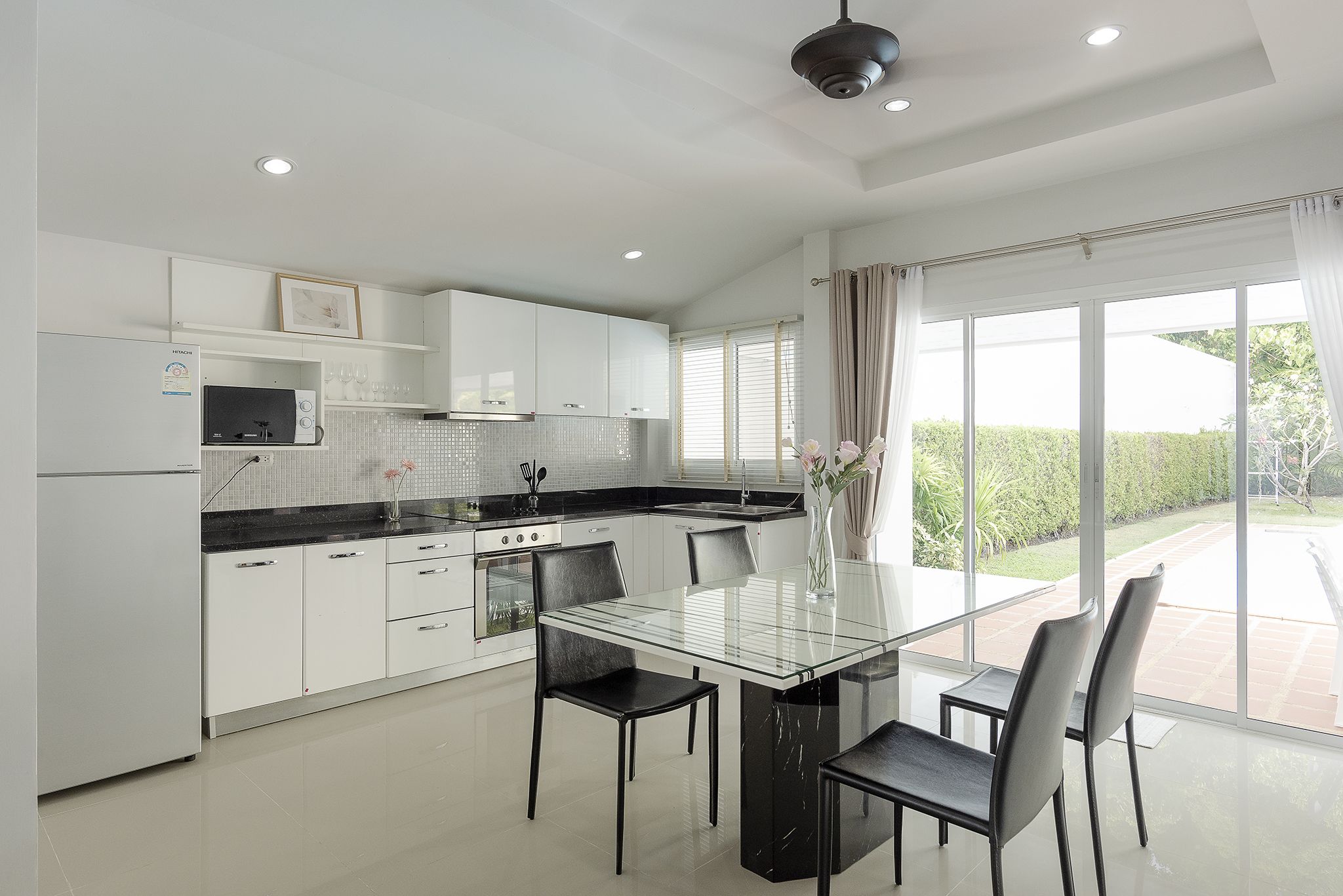 A modern kitchen with white cabinets, a glass dining table, and a view of the garden through large windows.