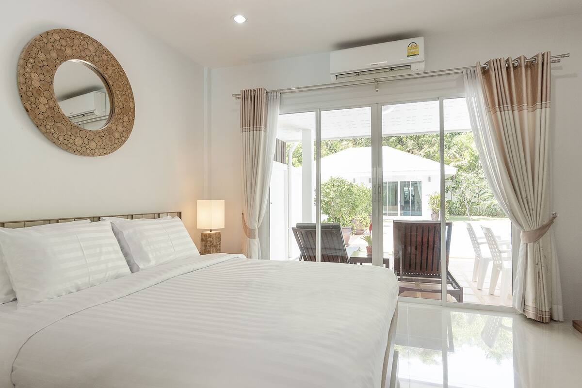 Une chambre d’hôtel avec un grand lit, un miroir rond, une fenêtre avec des rideaux et un appareil de climatisation.
