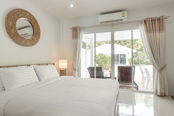 A hotel room with a large bed, a round mirror, a window with curtains, and an air conditioning unit. - BaanPromphun Pool Villa by BV (Pa Klok)