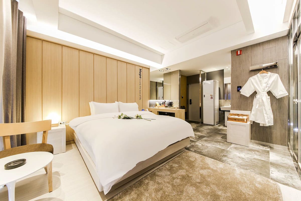 Ein modernes Hotelzimmer mit einem großen Bett, einem Nachttisch, einem Sessel, einem Kleiderschrank, einem Badezimmer mit Spiegel und einem an der Wand hängenden Bademantel.