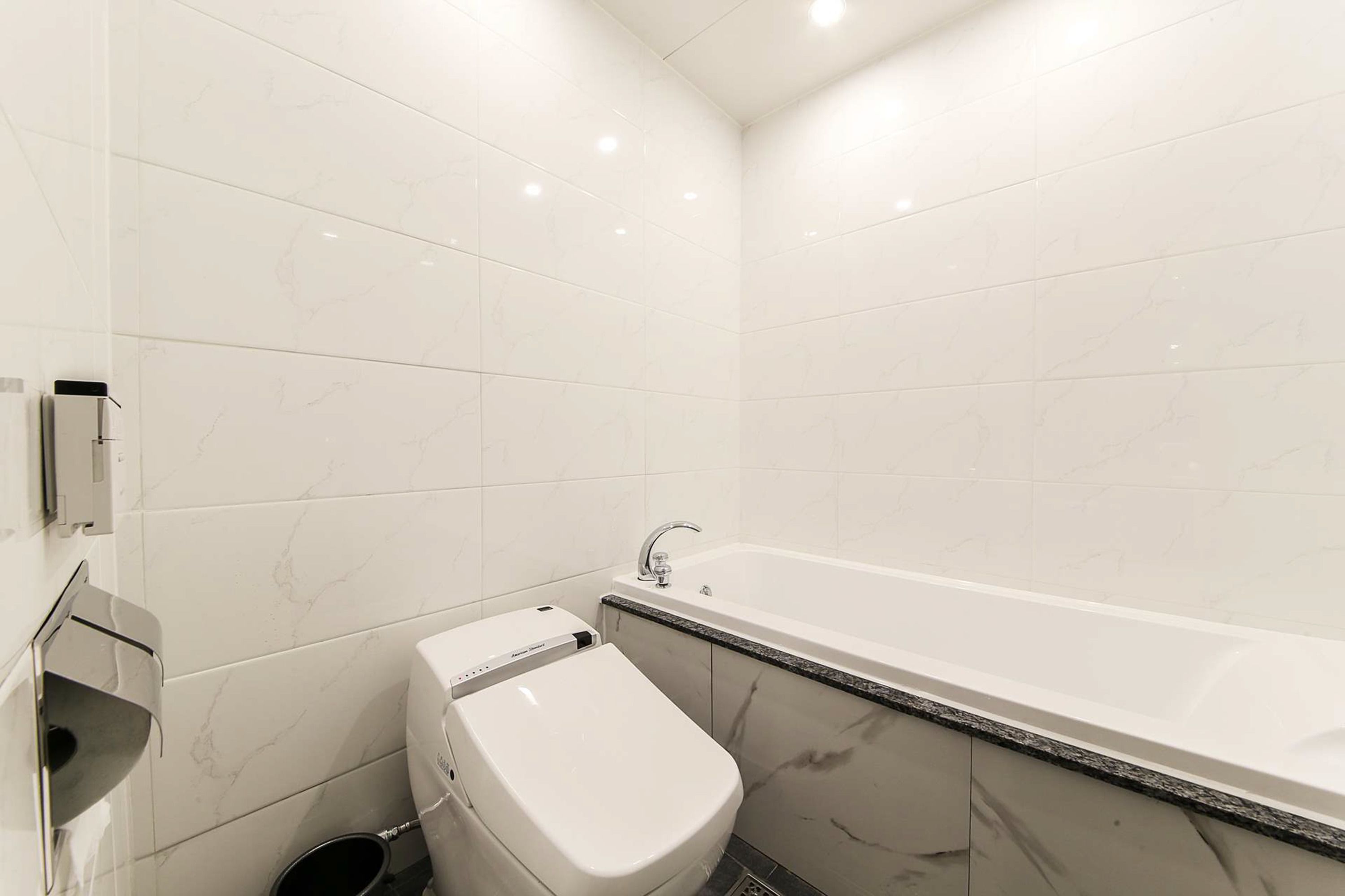 Un baño moderno con baldosas de mármol blanco, un inodoro blanco, una bañera con detalle negro y un secador de manos instalado en la pared.