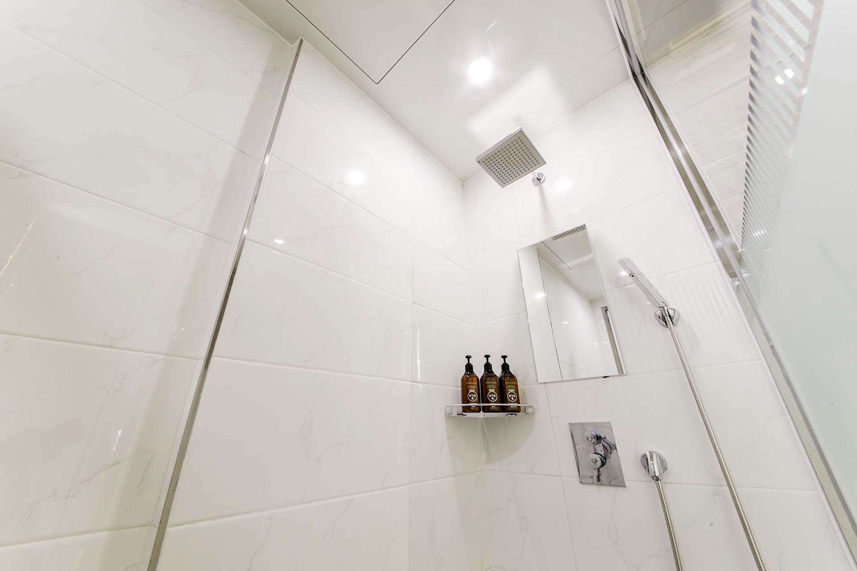 Un baño moderno con baldosas de mármol blanco, una cabina de ducha de vidrio, un estante empotrado con artículos de aseo y un espejo montado en la pared.