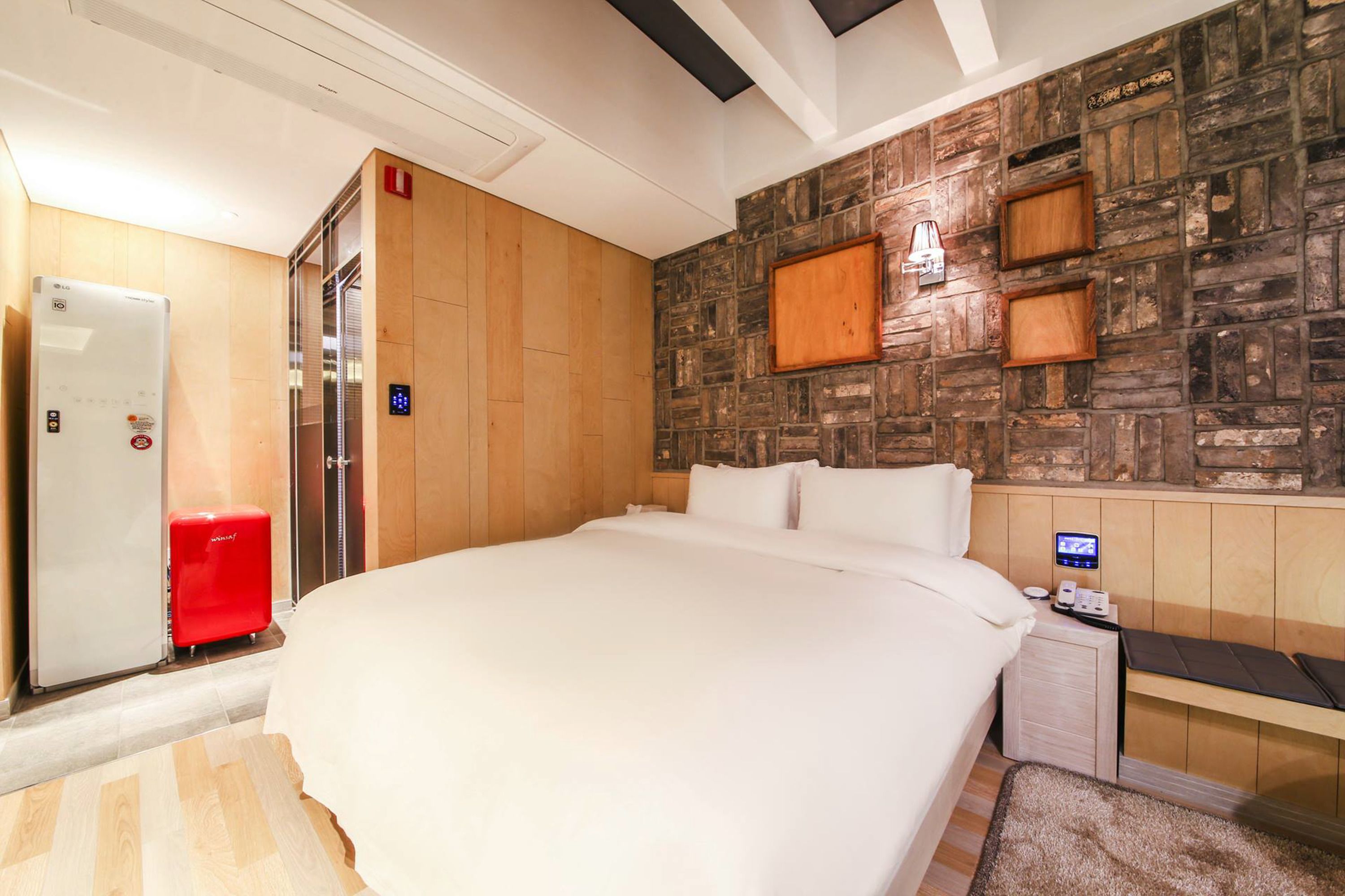 Habitación de hotel con una cama grande, un panel de pared de madera, una maleta roja, un minibar y un escritorio pequeño con una lámpara.