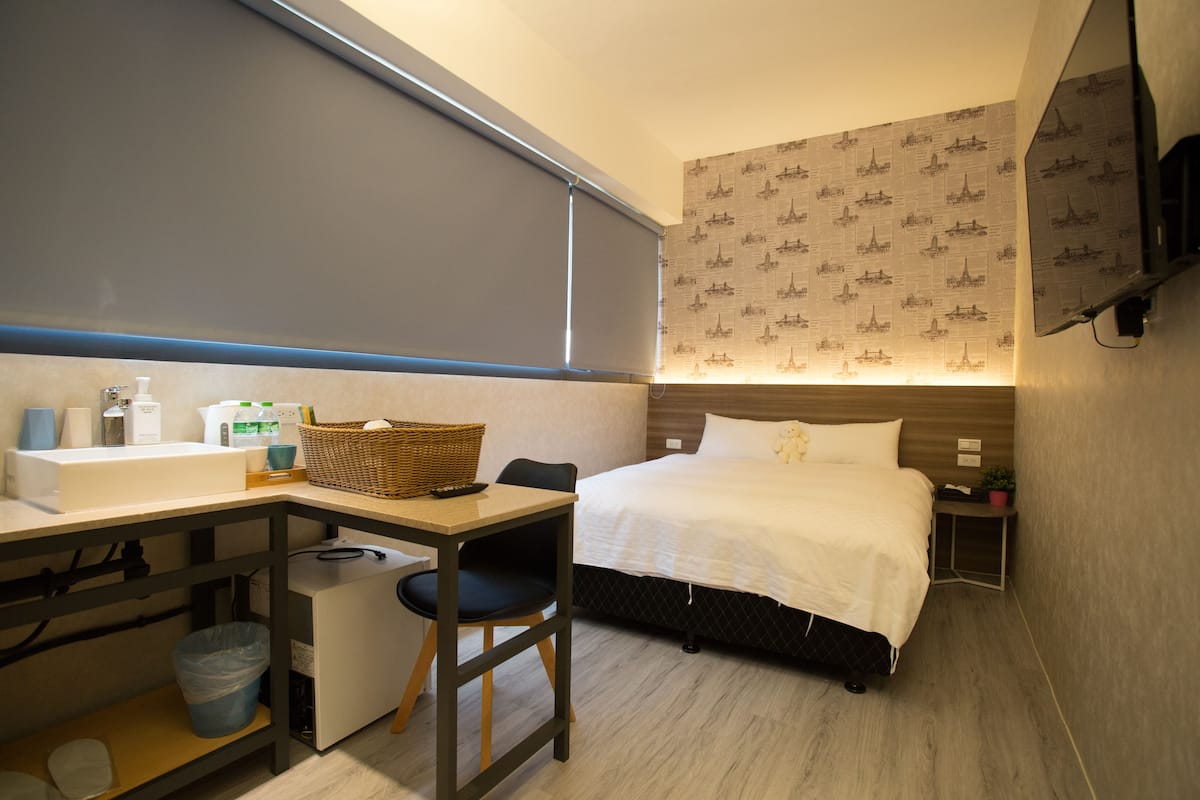 A hotel room with a bed, a desk, a chair, a small table, a basket, a trash bin, and a wall-mounted TV.