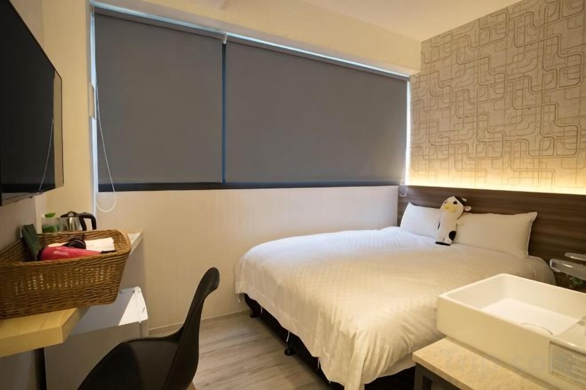 Una habitación de hotel con una cama, un escritorio con silla, una mesita de noche, un televisor montado en la pared y un diseño de pared estampado.