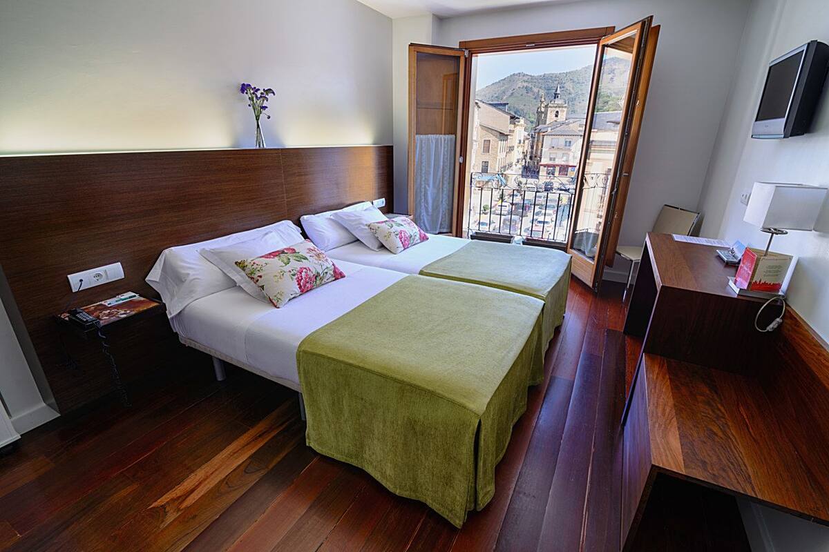 Habitación de hotel con dos camas, cabecera de madera, balcón con vistas a edificios, televisor y escritorio con lámpara.