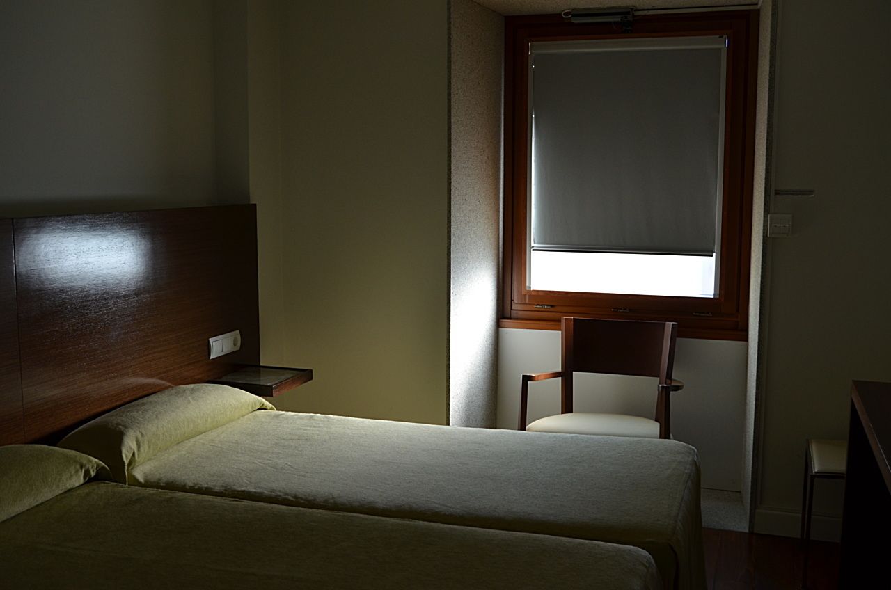 Una habitación pequeña y poco iluminada con dos camas, un cabecero de madera, una ventana con persiana y una silla.