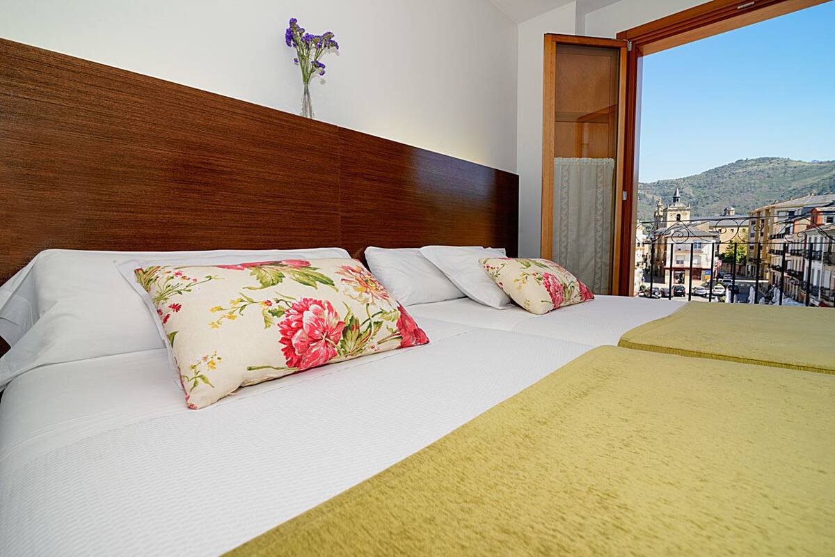 Habitación de hotel con cabecera de madera, dos camas con almohadas estampadas, vistas de la ciudad desde un balcón y un jarrón con flores en la pared.