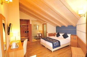 A hotel room with a wooden ceiling, a bed with a patterned bedspread, a desk with a chair, and a small table with a lamp. - Hotel B&B Bondi (Livigno)