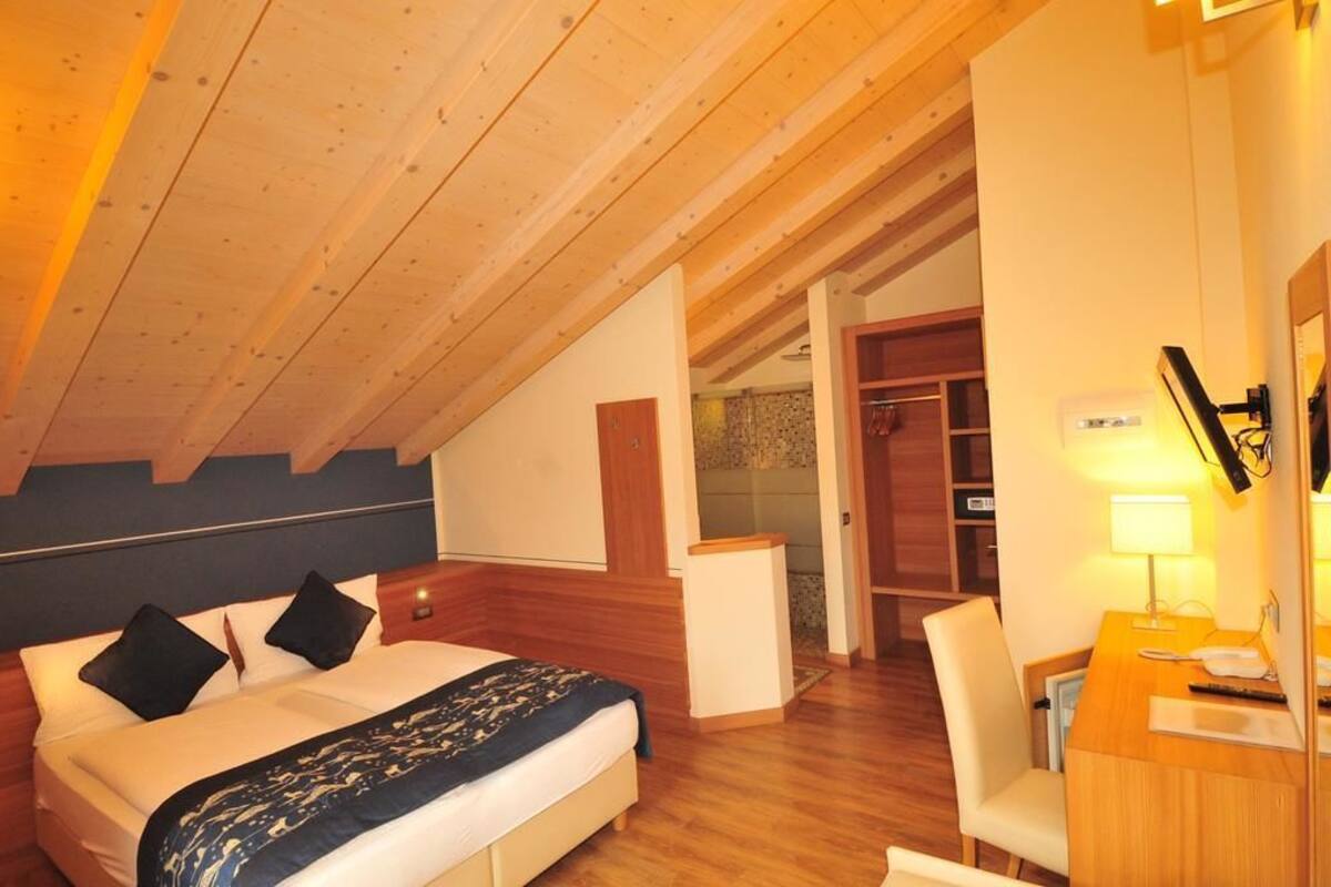A hotel room with a wooden ceiling, a bed with a patterned blanket, a desk with a lamp, and a bathroom visible through an open door.