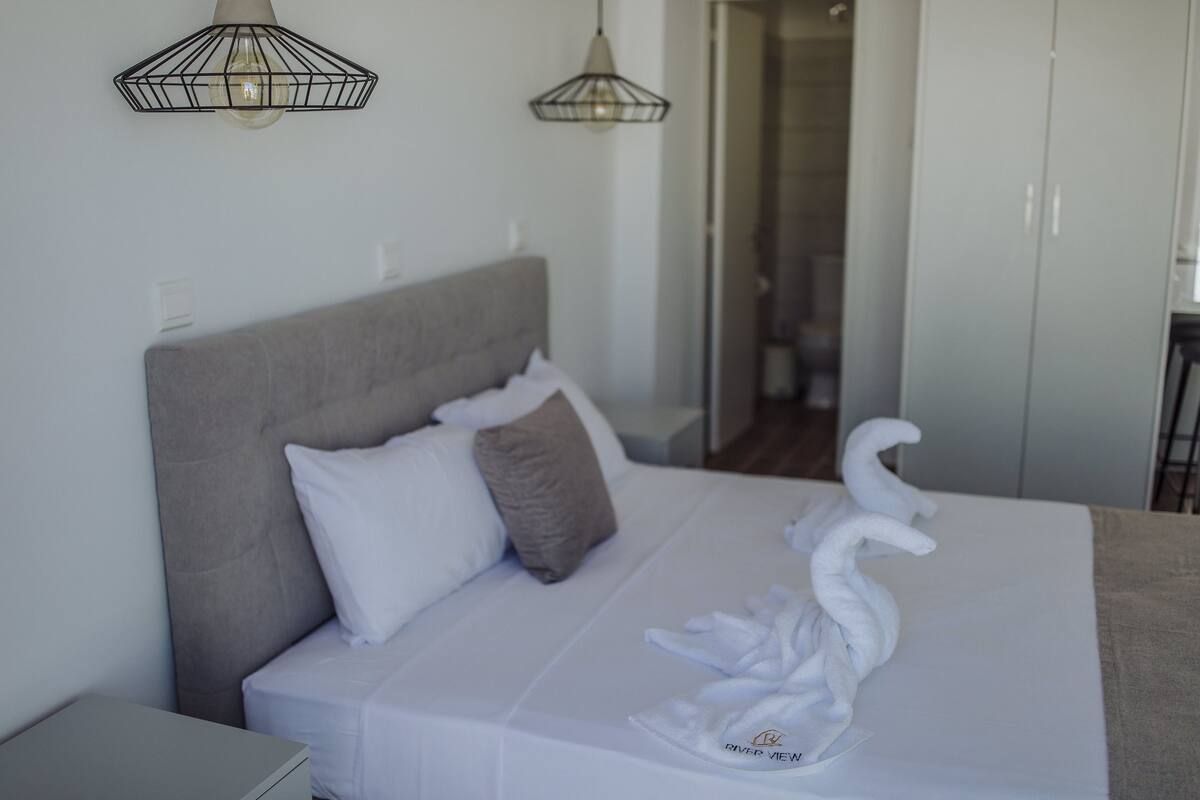 Una cama bien hecha con sábanas blancas, cabecero gris y una toalla con forma de cisne sobre la cama. Se ven dos lámparas colgantes sobre la cama, y un armario es visible al fondo.