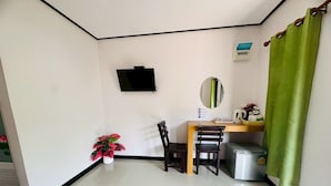 A small, clean room with a desk, chairs, a TV, a mirror, a small trash bin, and a potted plant. - Kanyaphat Gardenview Resort (Ban Dung)