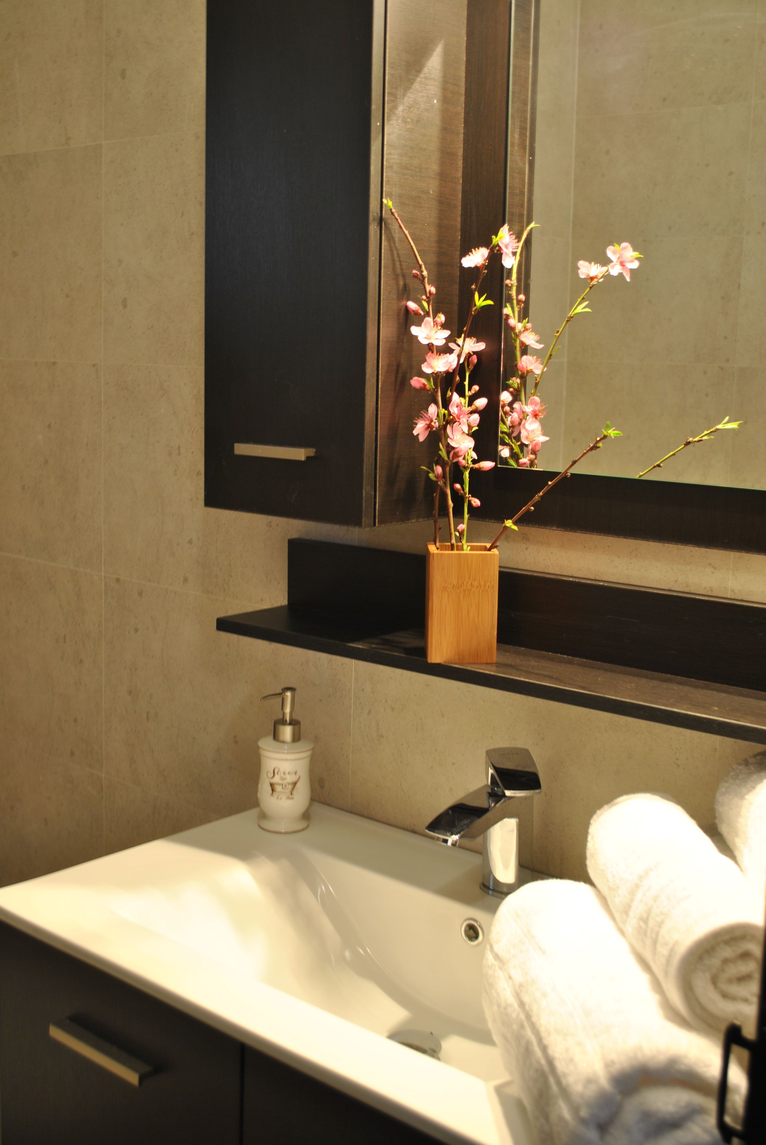 Un baño con un lavamanos blanco, un florero de madera con flores rosadas, un dispensador de jabón y un perchero con toallas.
