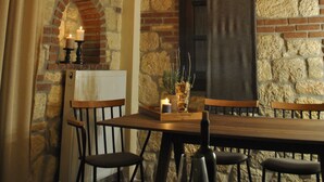 A stone-walled room with a wooden table and chairs, a hanging light fixture, and a brick fireplace.