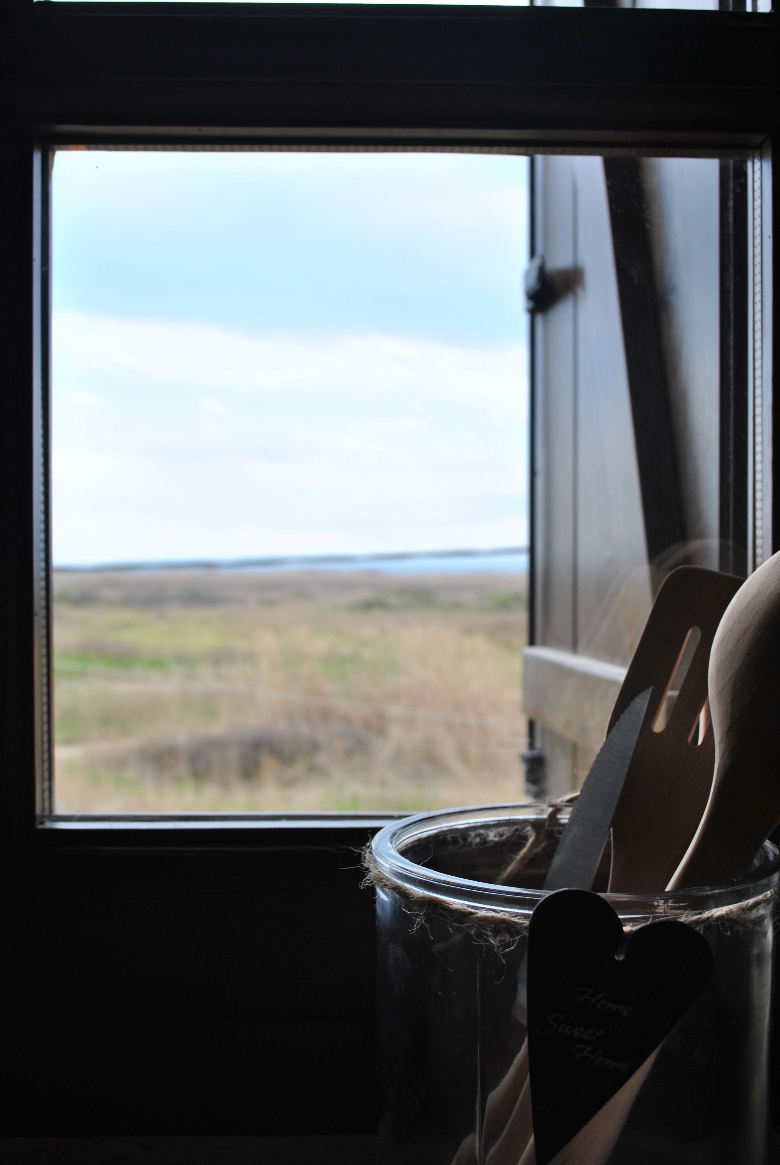 Uitzicht vanuit een raam op een begroeiend landschap met een eenvoudig interieur, inclusief een houten stoel en een metalen bak.