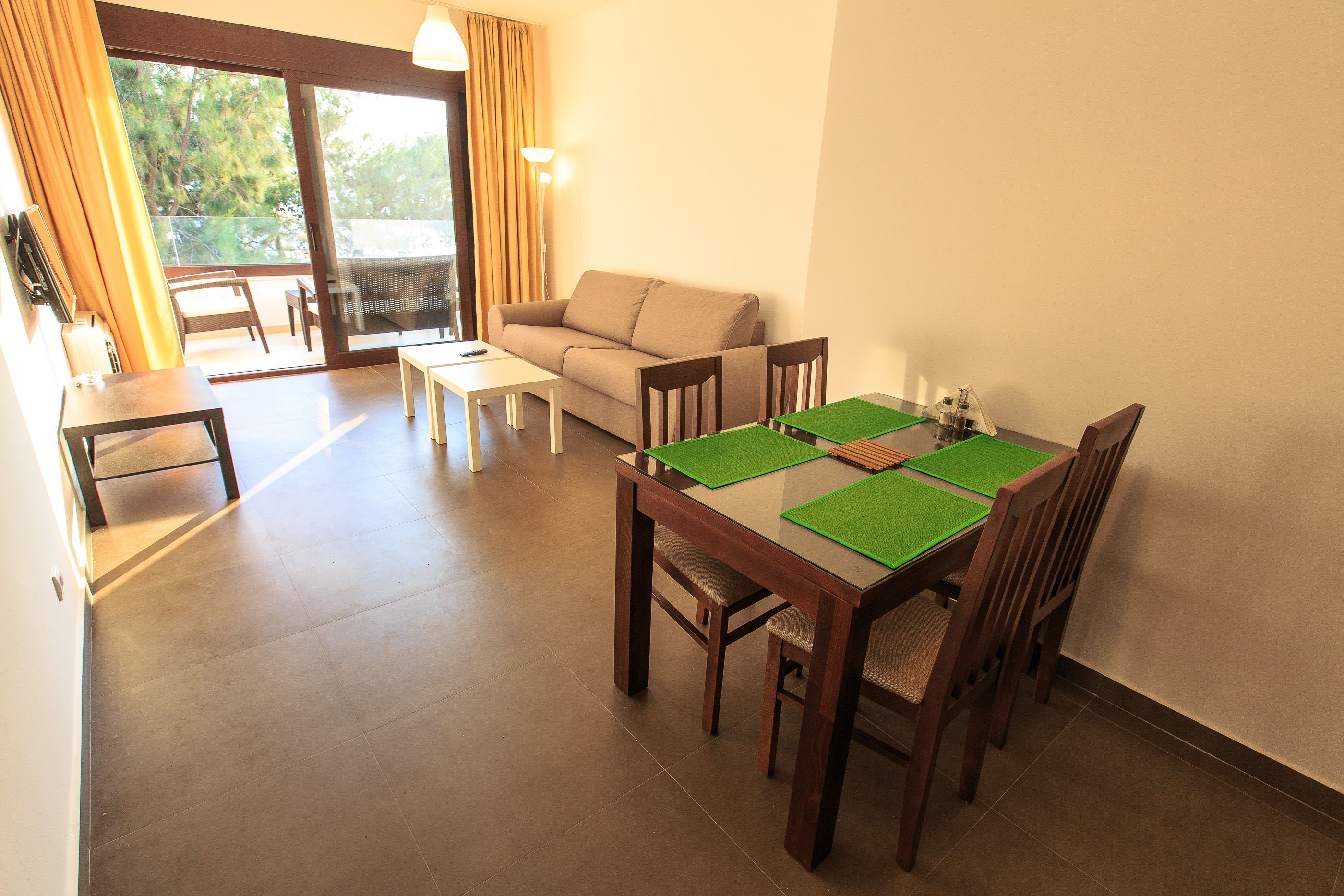 A dining area with a wooden table and chairs, a sofa, and a balcony with a view of trees.