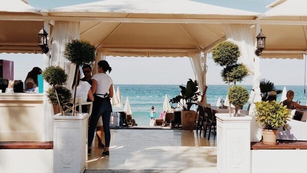 A seaside restaurant with outdoor seating, a clear view of the ocean, and a paved walkway leading to the entrance. - Apartments Kuce Lekovica snack bar- Bistro & Beach  (Bar)