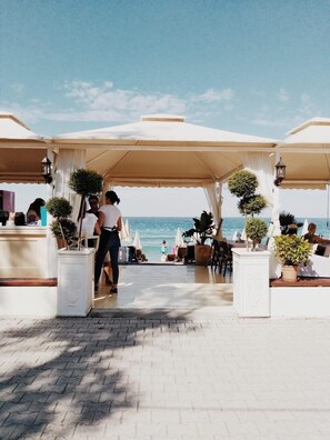 Un restaurant en bord de mer avec une terrasse, une vue dégagée sur l’océan et un chemin pavé menant à l’entrée.