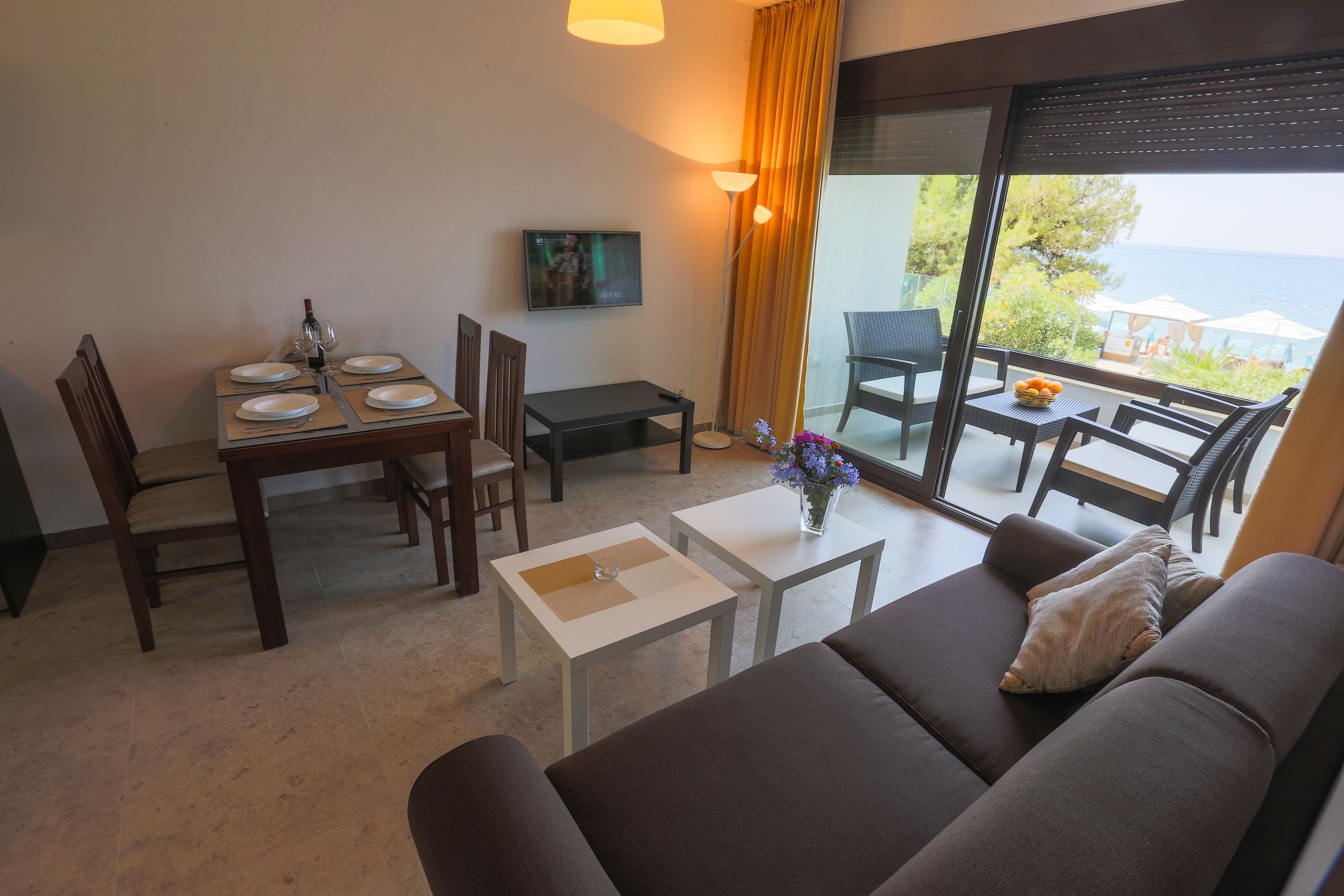 Un salon moderne avec un canapé, une table à manger et des chaises. Un téléviseur est fixé au mur, et un balcon offre une vue sur la mer.