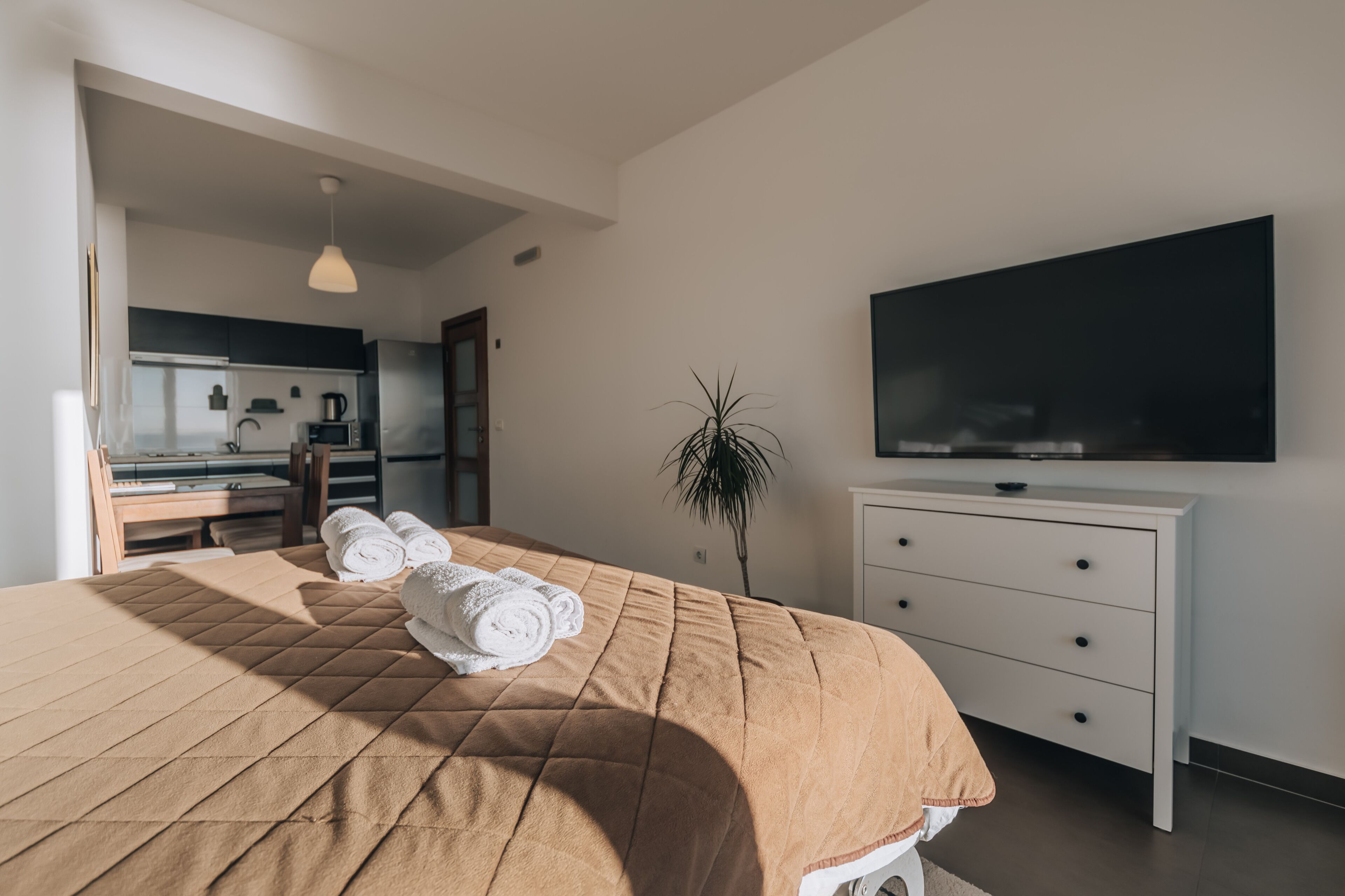 A modern hotel room with a large bed, a flat-screen TV, a white dresser, and a kitchenette in the background.