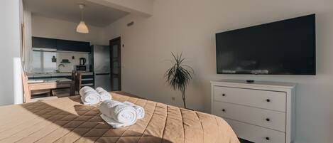 A modern hotel room with a large bed, a flat-screen TV, a white dresser, and a kitchenette in the background.