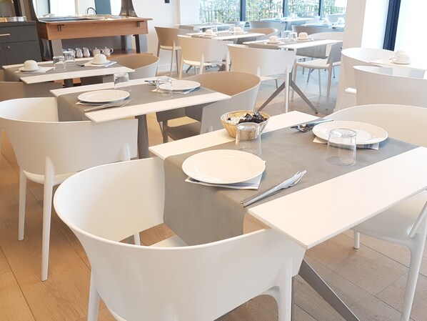 A modern dining area with white tables and chairs, neatly arranged with plates, glasses, and cutlery.