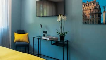 A modern hotel room with a bed, a desk with a lamp, a chair, a potted plant, and a painting of a building.