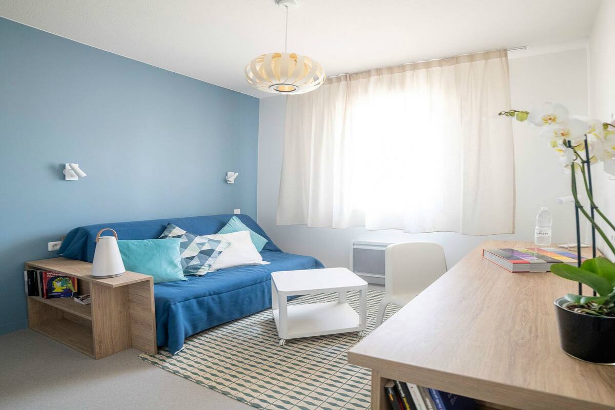 Et moderne soveværelse med blå vægge, en seng med dynebetræk i blå og hvid, et natbord med lampe, et lille skrivebord med stol, en orkidé i potte og et vindue med gardiner.