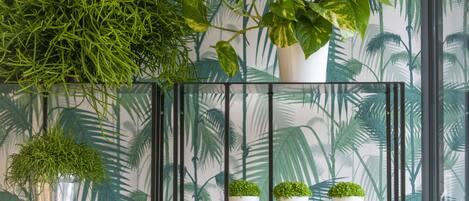 Una habitación con papel tapiz tropical, un estante de metal con plantas en macetas y una ventana con vistas a palmeras.