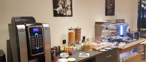 A modern coffee station with a Krea espresso machine, various coffee pods, and a display of pastries and breakfast items.