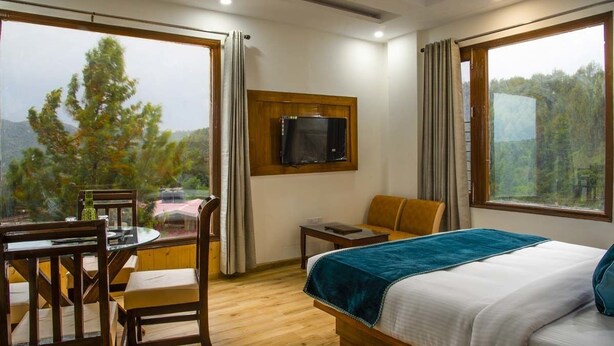 A hotel room with a bed, a TV mounted on the wall, a dining table with chairs, and a view of the outdoors. - Jade Vine Resort (Shimla)