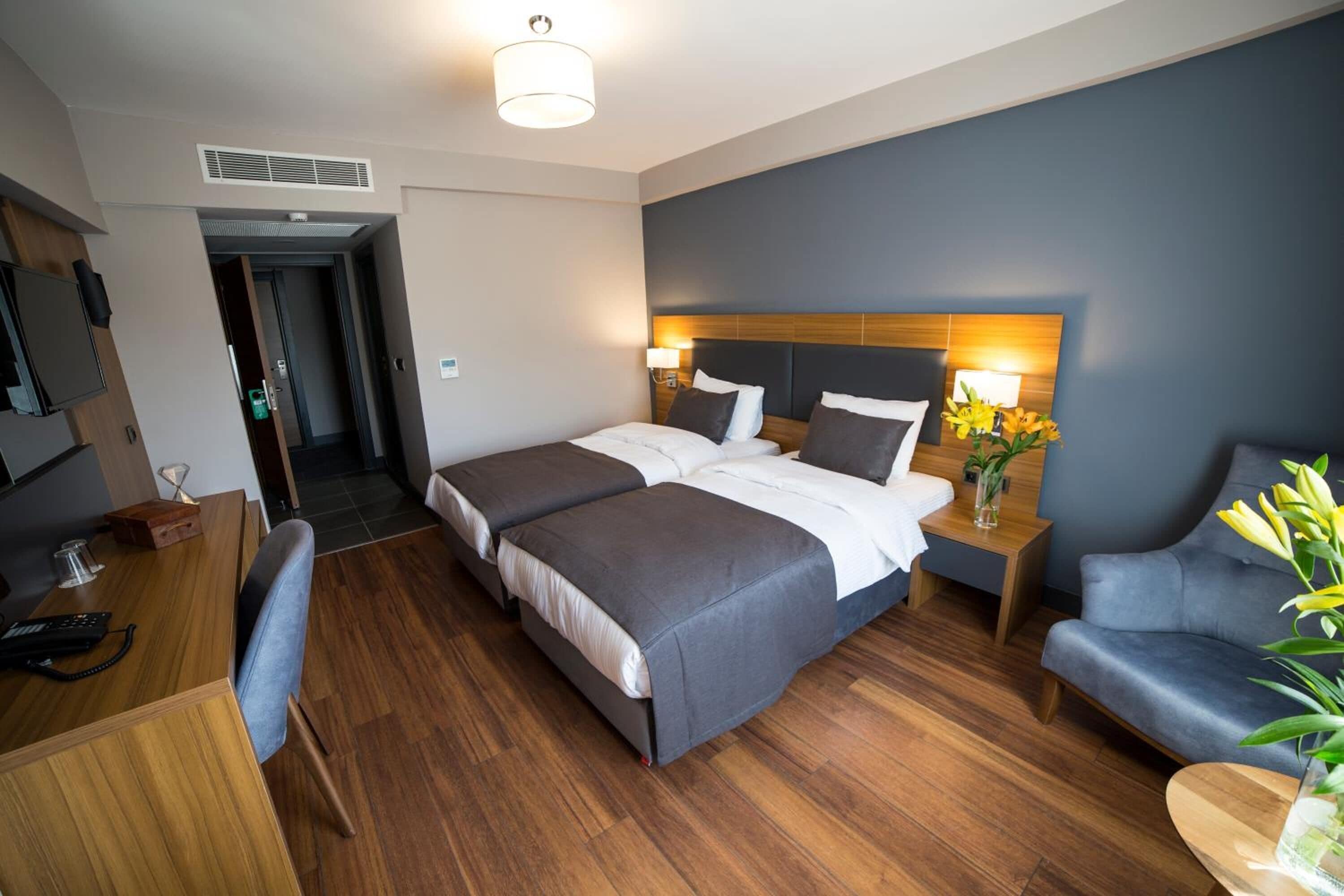 A hotel room with a large bed, a desk, a chair, and a bedside table with a lamp and flowers.