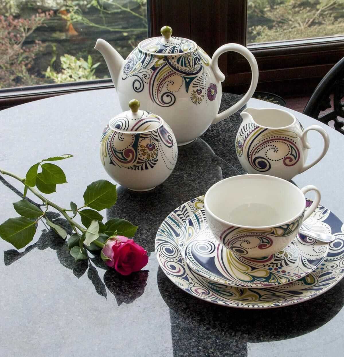 Ein gedeckter Tisch mit Teekanne, Tasse und Untertasse, verziert mit aufwendigen Mustern. Eine Rose steht neben dem Tisch.