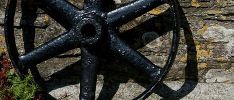 A black metal wheel against a stone wall with a garden below featuring various flowers.