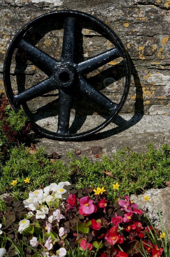 Una rueda de metal negro contra una pared de piedra, con un jardín inferior que muestra diversas flores.