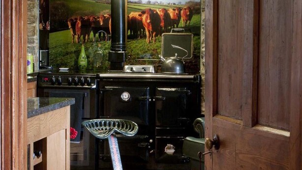 A rustic kitchen with a stone wall, wooden shelves, and a large stove. - Tregondale Manor Farm (Liskeard)