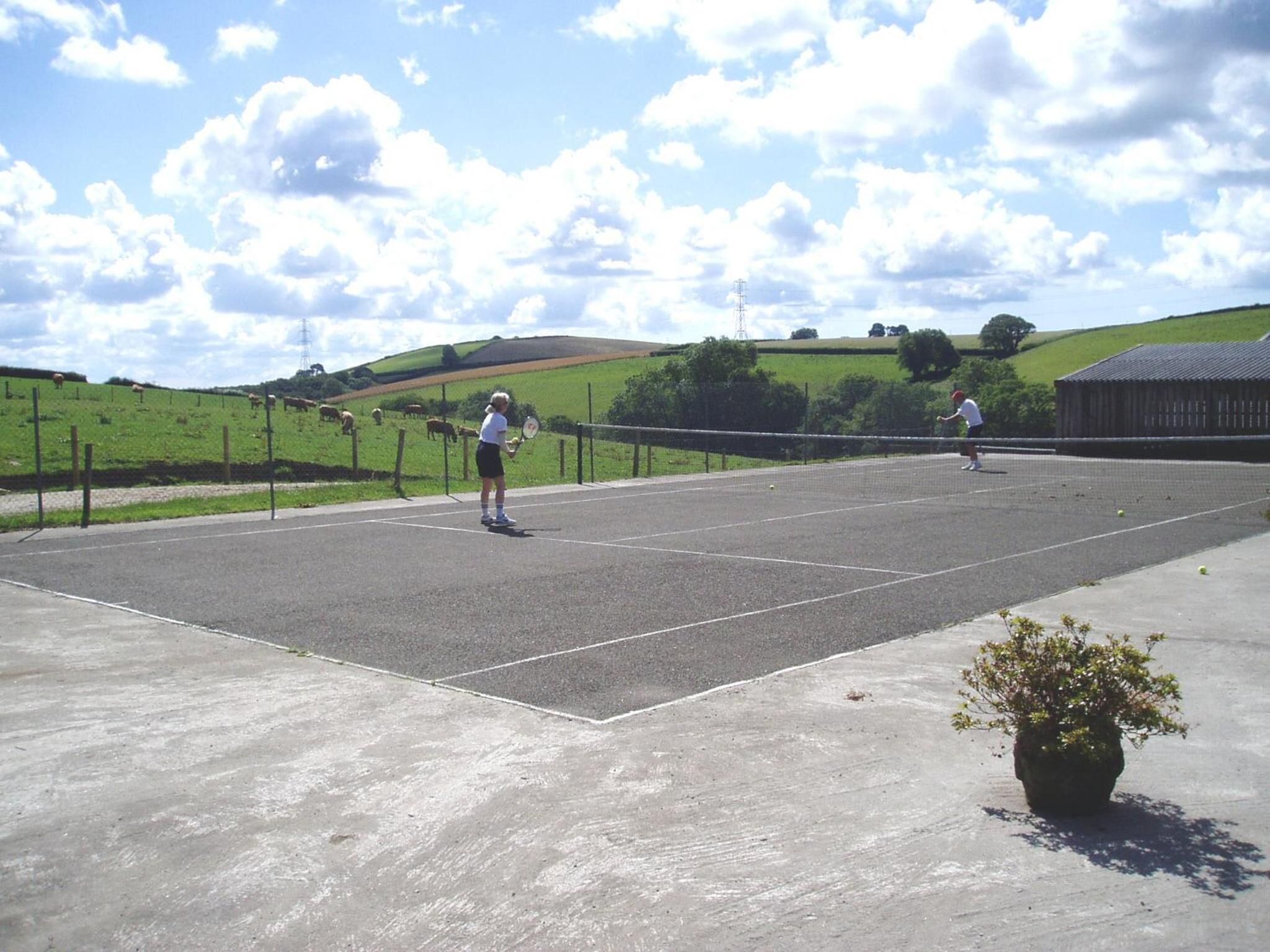 Cancha de tenis al aire libre con dos jugadores, rodeada de campos verdes y colinas.