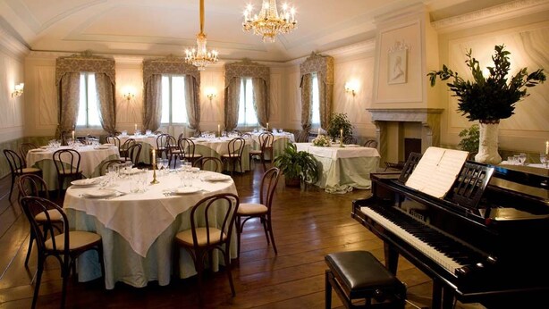 Une salle à manger élégante, avec des tables rondes, des chaises, des lustres et un grand piano.