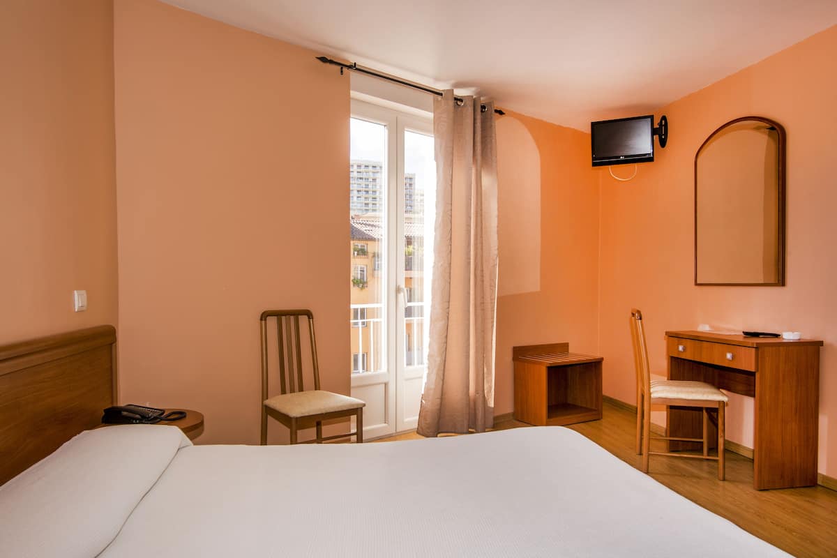 Quarto de hotel com cama, escrivaninha, cadeira, televisão e janela com cortinas.