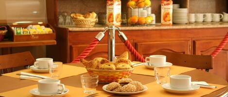 Desayuno con una mesa para dos, incluyendo croissants, una cesta de pasteles y un vaso de agua.