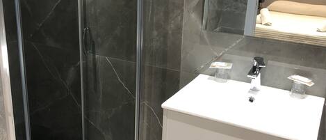 A modern bathroom with a glass-enclosed shower, a white vanity with a mirror, and a bed visible in the background.