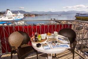 A balcony with wicker chairs, a table with breakfast items, and a view of ships and mountains. - Le Dauphin Hotel (Ajaccio)