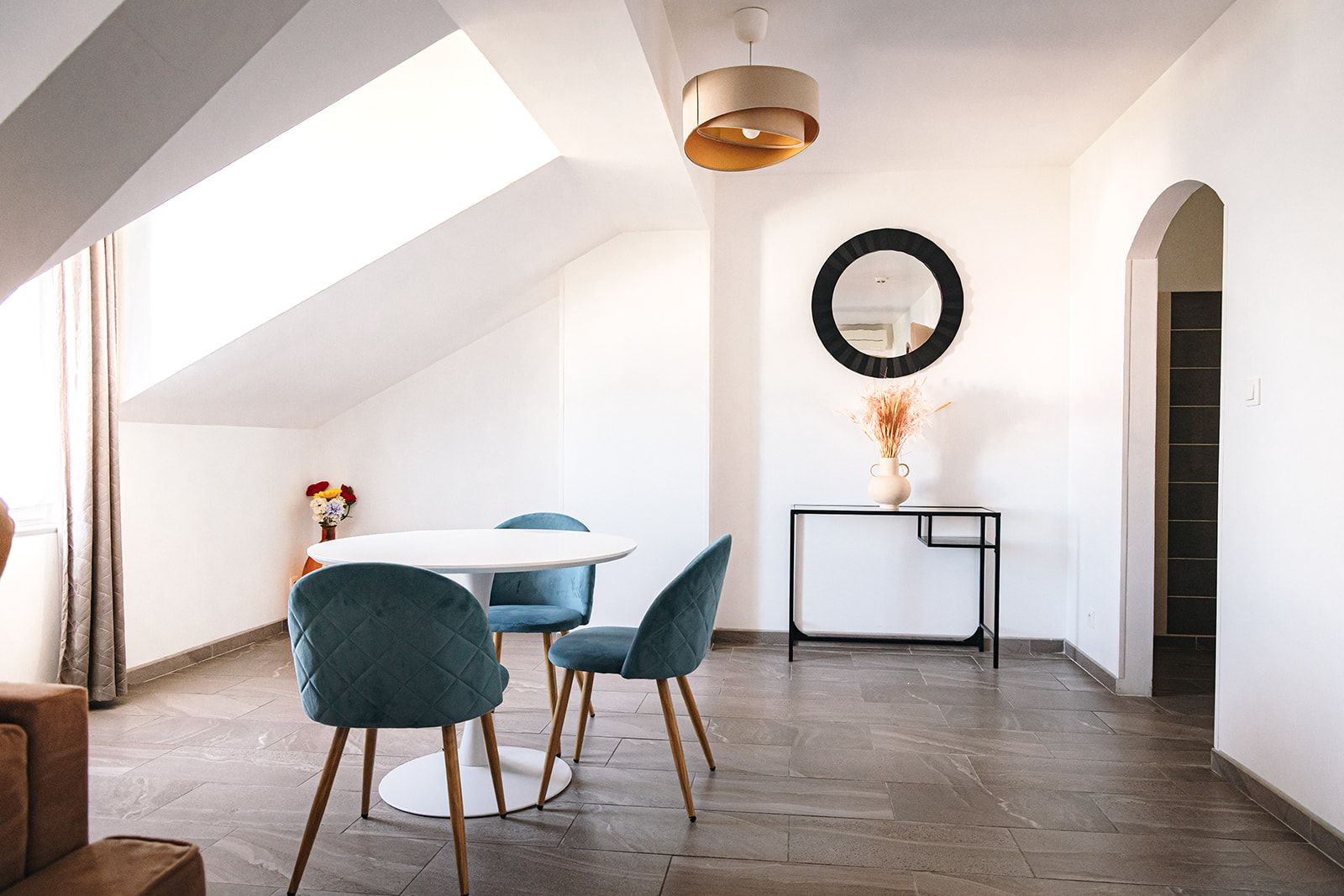 A modern interior with a round dining table, blue upholstered chairs, a black console table with a vase, and a large mirror on the wall.
