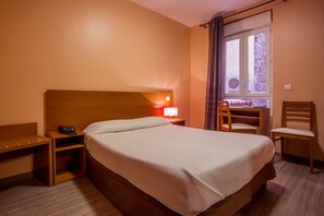 Ett hotellrum med en säng, ett skrivbord, en stol och ett fönster med gardiner.