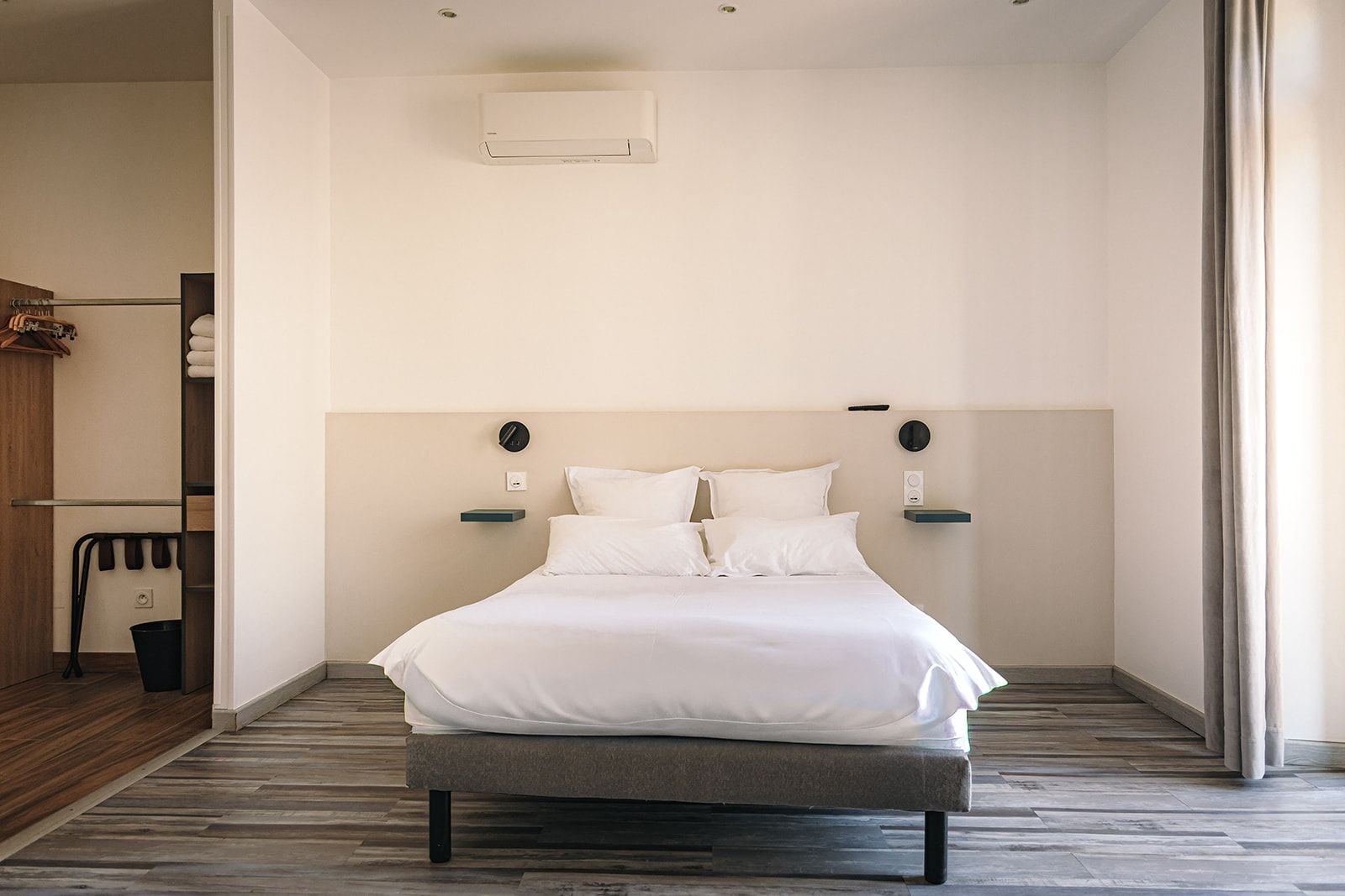 Una habitación de hotel moderna con una cama grande, una mesita de noche pequeña, aire acondicionado instalado en la pared y suelo de madera.