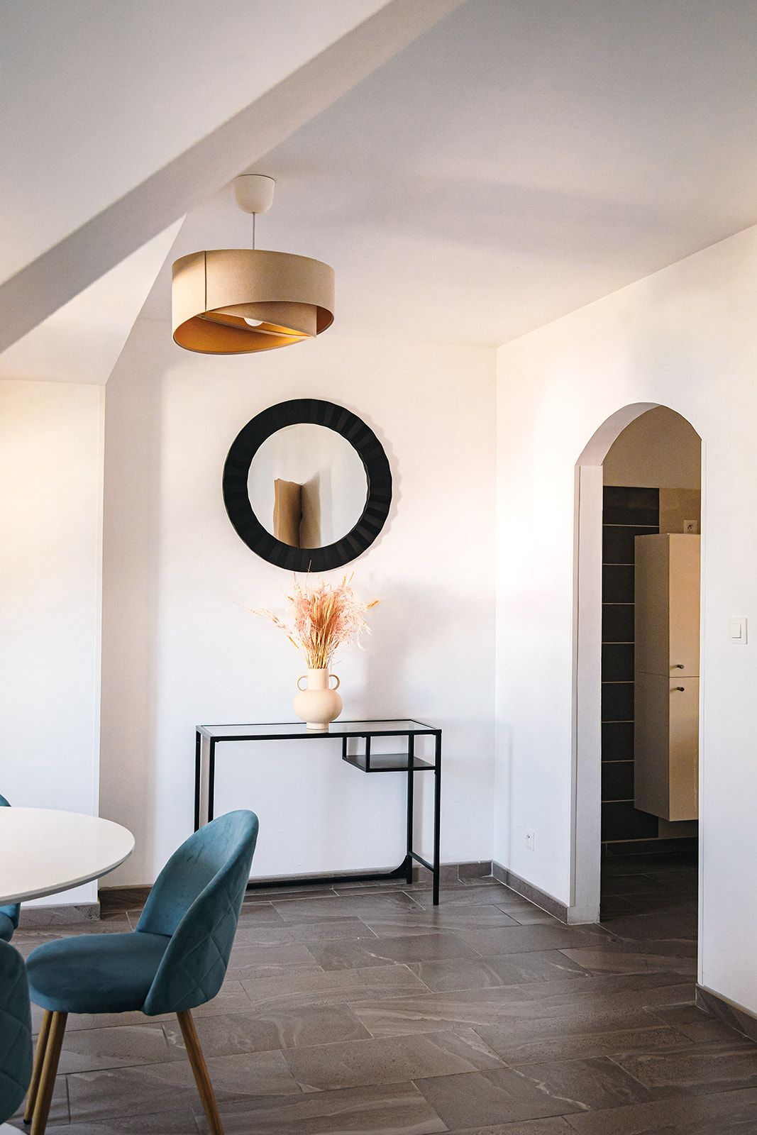 A modern interior with a dining area, a round mirror, a vase with dried plants, and a staircase leading to another level.