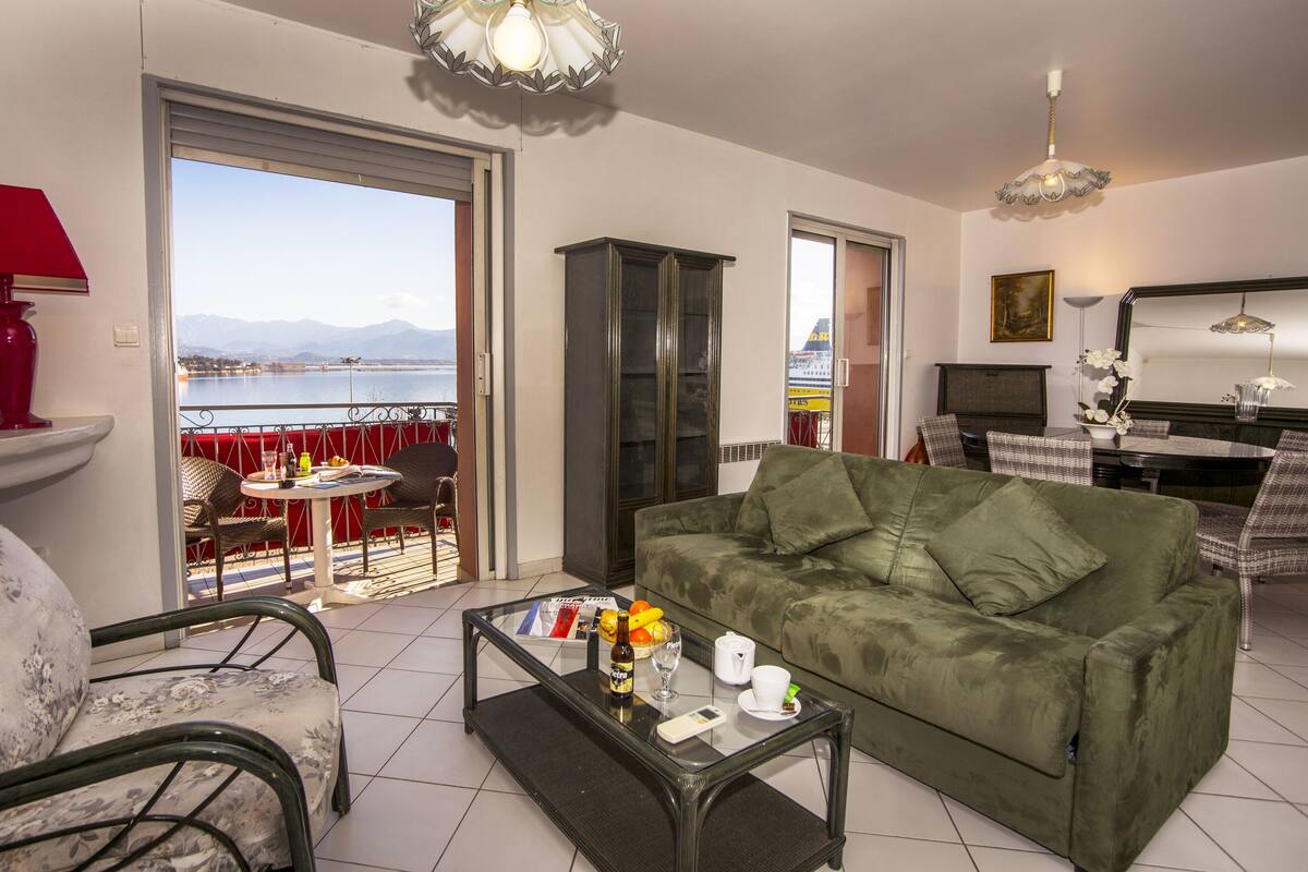 Uma sala de estar com um sofá verde, uma mesa de centro de vidro com uma garrafa, copos e um controle remoto, uma área de jantar com mesa e cadeiras, e vista para um corpo de água e montanhas por uma grande janela.